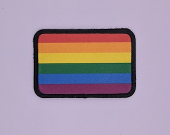 Pode incluir: Um remendo de bandeira do orgulho arco-íris com listras vermelhas, laranja, amarelas, verdes, azuis e roxas. O remendo é cercado por uma borda preta.