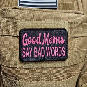 May include: A black patch with white stitching features the text "Good Moms Say Bad Words" in pink lettering. The patch is attached to a brown tactical backpack.