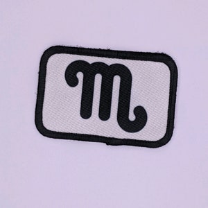 Puede incluir: Un parche rectangular con fondo blanco y borde negro. El parche presenta una letra "m" negra estilizada en el centro. El parche es probablemente para ropa o accesorios.