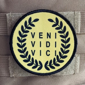 Pode incluir: Um remendo circular amarelo com uma coroa de louro preta e o texto "VENI VIDI VICI" em preto.