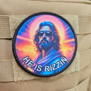 May include: A round, black-bordered patch featuring a colourful image of a man with sunglasses and a beard. The background is a gradient of pink, orange, and blue. The text "HE IS RIZZIN" is printed in white on the patch.