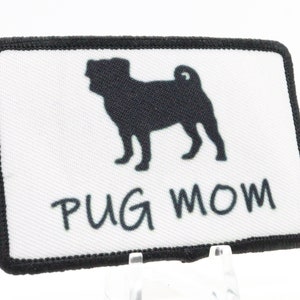 Puede incluir: Un parche rectangular blanco con costuras negras alrededor de los bordes. Una silueta negra de un carlino está centrada en el parche. Debajo del carlino está el texto "PUG MOM" en negro.