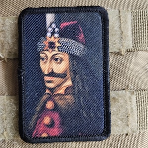 Può includere: Una toppa rettangolare con un ritratto di una figura storica con i baffi, che indossa un cappello e abiti ornati. La toppa ha un bordo nero ed è posta su uno sfondo beige.