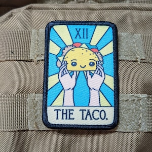 以下が含まれることがあります： 笑顔の顔をした漫画のタコスが描かれた黒と黄色のパッチ。パッチには「XII THE TACO.」という文字が書かれています。