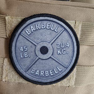 Op de afbeelding: Een ronde, grijze patch met het woord "BARBELL" in een boog aan de boven- en onderkant. Het midden toont een halter gewichtplaat ontwerp, met "45 LB." en "20,4 KG." erop gedrukt. De patch is genaaid op een bruin textiel.