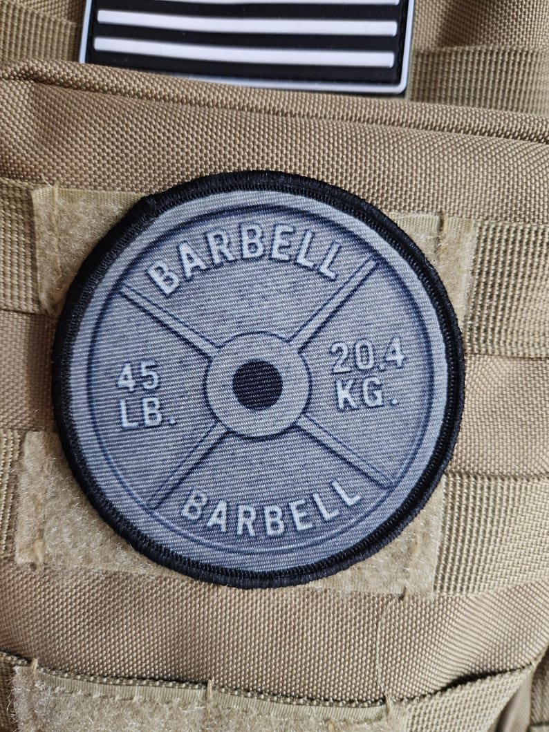 May include: A round, gray patch with the word "BARBELL" arched at the top and bottom. The center features a barbell weight plate design, with "45 LB." and "20.4 KG." printed on it. The patch is sewn onto a tan fabric.