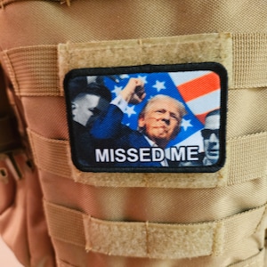 May include: A tan tactical vest with a black patch featuring a photo of a man with a red, white, and blue American flag in the background. The text on the patch reads "MISSED ME".
