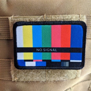 Peut inclure: Un patch noir et blanc "NO SIGNAL" avec un motif d'essai de télévision coloré. Le patch est fixé à un fond en tissu brun.