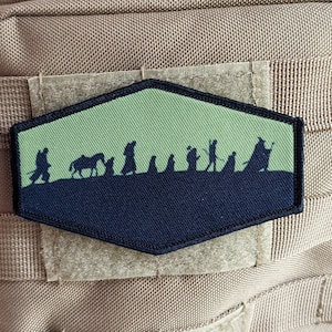 May include: A green and black embroidered patch with a silhouette of a group of figures walking across a landscape. The figures are in a line and appear to be walking towards the right side of the patch. The patch is shaped like a hexagon and has a dark blue border.