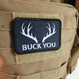 Puede incluir: Un parche negro con cuernos blancos y el texto "BUCK YOU" sobre un fondo marrón.