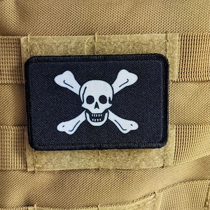 May include: A black and white skull and crossbones patch with a white background. The patch is sewn onto a tan fabric.