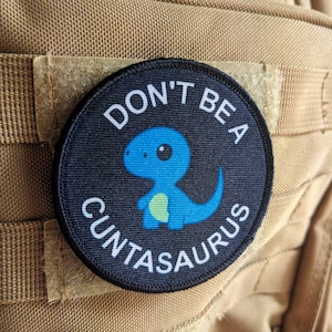 Puede incluir: Un parche blanco y negro con un dinosaurio azul y el texto "Don't be a Cuntasaurus".