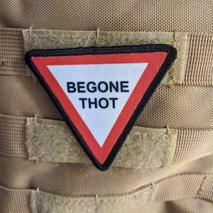 May include: A red and white triangular warning sign with black trim and the text "BEGONE THOT" in black lettering. The sign is attached to a brown fabric background.
