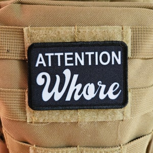 Può includere: Una toppa nera con testo bianco che recita "ATTENTION Whore" attaccata a un giubbotto tattico beige.