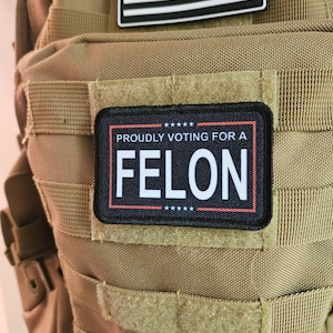 Puede incluir: Una mochila táctica de color marrón claro con un parche negro que dice "Proudly Voting For A Felon" con cinco estrellas arriba y abajo del texto. El parche está sujeto con velcro. Hay un parche más pequeño de la bandera estadounidense por encima del parche más grande.