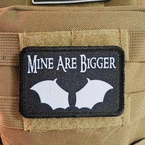 Pode incluir: Um remendo preto com costuras brancas e o texto "MINE ARE BIGGER" em letras brancas. O remendo apresenta duas asas de morcego brancas.