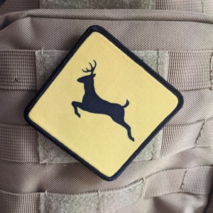 May include: A yellow square patch with a black silhouette of a deer jumping. The patch is sewn onto a brown fabric background.