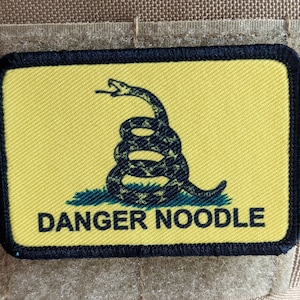 Pode incluir: Um remendo amarelo com uma cobra preta enrolada em torno de um pau e o texto "DANGER NOODLE".