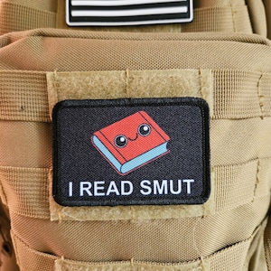 May include: A tan tactical backpack with a black and white American flag patch and a black patch with a red book with a smiley face and the text "I READ SMUT".