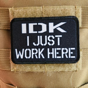 Könnte beinhalten: Schwarzer Patch mit weißem Text, der "IDK I JUST WORK HERE" auf einem braunen Hintergrund lautet.
