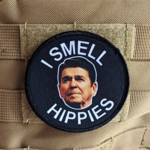 May include: A black and white round patch with a picture of George W. Bush and the text "I Smell Hippies" in white.