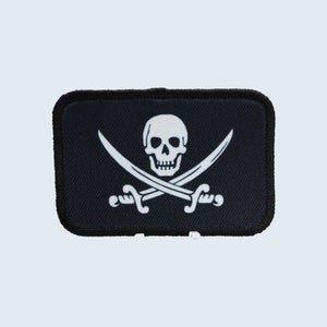 Puede incluir: Un parche de tela negro con un diseño de calavera y huesos cruzados blancos. La calavera tiene dos cuencas de ojos y una boca. Los huesos cruzados están hechos de dos espadas.