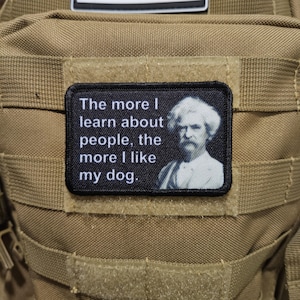 Könnte beinhalten: Ein rechteckiger schwarzer Aufnäher mit dem Zitat "Je mehr ich über Menschen lerne, desto mehr mag ich meinen Hund." Der Aufnäher zeigt ein Schwarz-Weiß-Porträt eines Mannes. Der Aufnäher ist an einem beigefarbenen Rucksack befestigt.