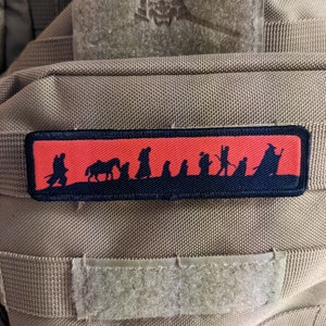 Pode incluir: Um patch retangular com um design de silhueta preta sobre um fundo vermelho. O design retrata figuras e animais. O patch é costurado em um tecido bege com fecho de velcro.