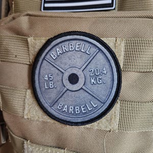 May include: A round patch with the word "BARBELL" in a semi-circle at the top and bottom. The patch is gray with black trim and features the weight "45 LB." and "20.4 KG." on the face. The patch is attached to a tan backpack.