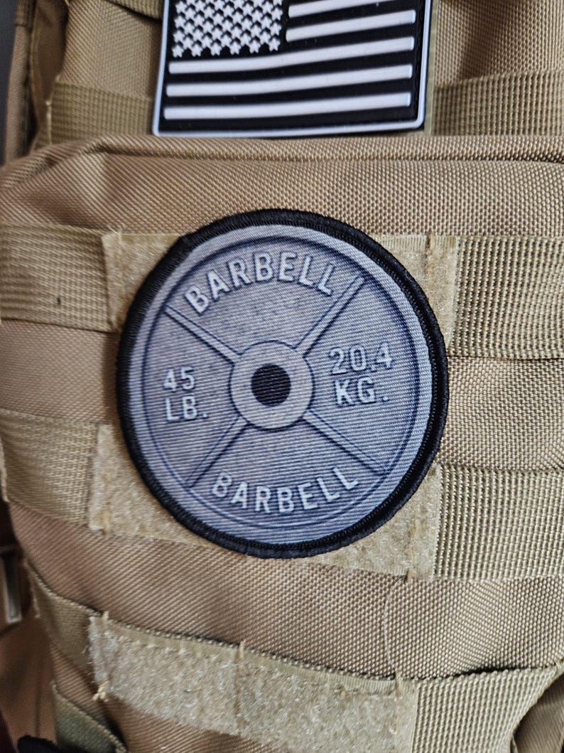 May include: A round patch with the word "BARBELL" in a semi-circle at the top and bottom. The patch is gray with black trim and features the weight "45 LB." and "20.4 KG." on the face. The patch is attached to a tan backpack.