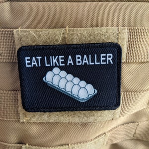 Pode incluir: Um remendo preto com texto branco que diz "EAT LIKE A BALLER" e uma ilustração branca de uma caixa de ovos.