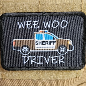 May include: Black and white patch with a cartoon sheriff's truck and the text "WEE WOO DRIVER".