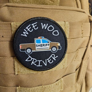 Puede incluir: Un parche blanco y negro con un camión de sheriff de dibujos animados y el texto "WEE WOO DRIVER" alrededor.