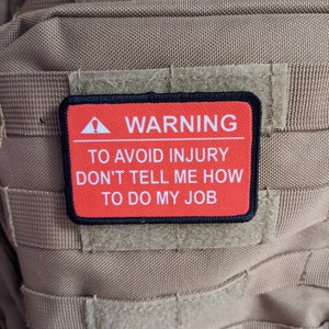 Op de afbeelding: Rode en zwarte waarschuwingsbadge met witte tekst die "WARNING TO AVOID INJURY DON'T TELL ME HOW TO DO MY JOB" luidt.