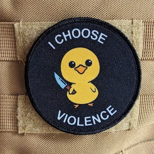 May include: A circular black patch featuring the text "I CHOOSE VIOLENCE" around a yellow cartoon duck holding a knife. The patch is attached to a tan fabric background.