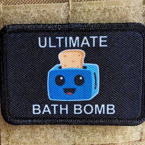 Könnte beinhalten: Schwarzer Patch mit weißem Text, der "ULTIMATE BATH BOMB" lautet, und einem Cartoon-Bild eines blauen Toasters mit einem lächelnden Gesicht und einer Scheibe Brot darin.