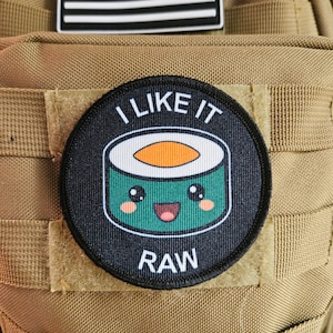 Puede incluir: Un parche redondo negro y verde con un rollo de sushi de dibujos animados con una cara sonriente. El texto "I LIKE IT RAW" está impreso en el parche.