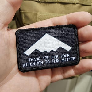 May include: A black rectangular patch with a white stealth bomber design and the text "THANK YOU FOR YOUR ATTENTION TO THIS MATTER" in white. The patch is held in a hand, with a tan background.