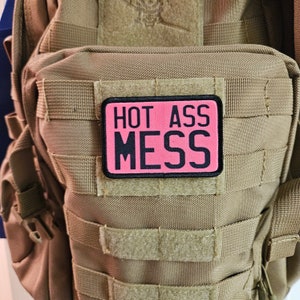 May include: A pink patch with black text that reads "HOT ASS MESS" is attached to a brown tactical backpack.