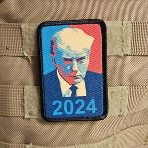 May include: A black and white patch with a red, white, and blue image of a man's face. The text "2024" is printed in blue on the patch.