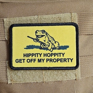 May include: A yellow and black patch with a cartoon frog holding a rifle. The text on the patch reads "Hippity Hoppity Get Off My Property".