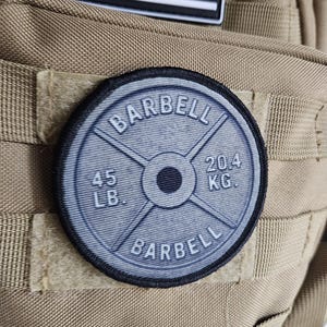 May include: A round, gray barbell weight patch with the word "BARBELL" in a circular pattern. The patch displays "45 LB" and "20.4 KG" on the weight. The patch is attached to a tan tactical backpack, with a black and white American flag patch above it.
