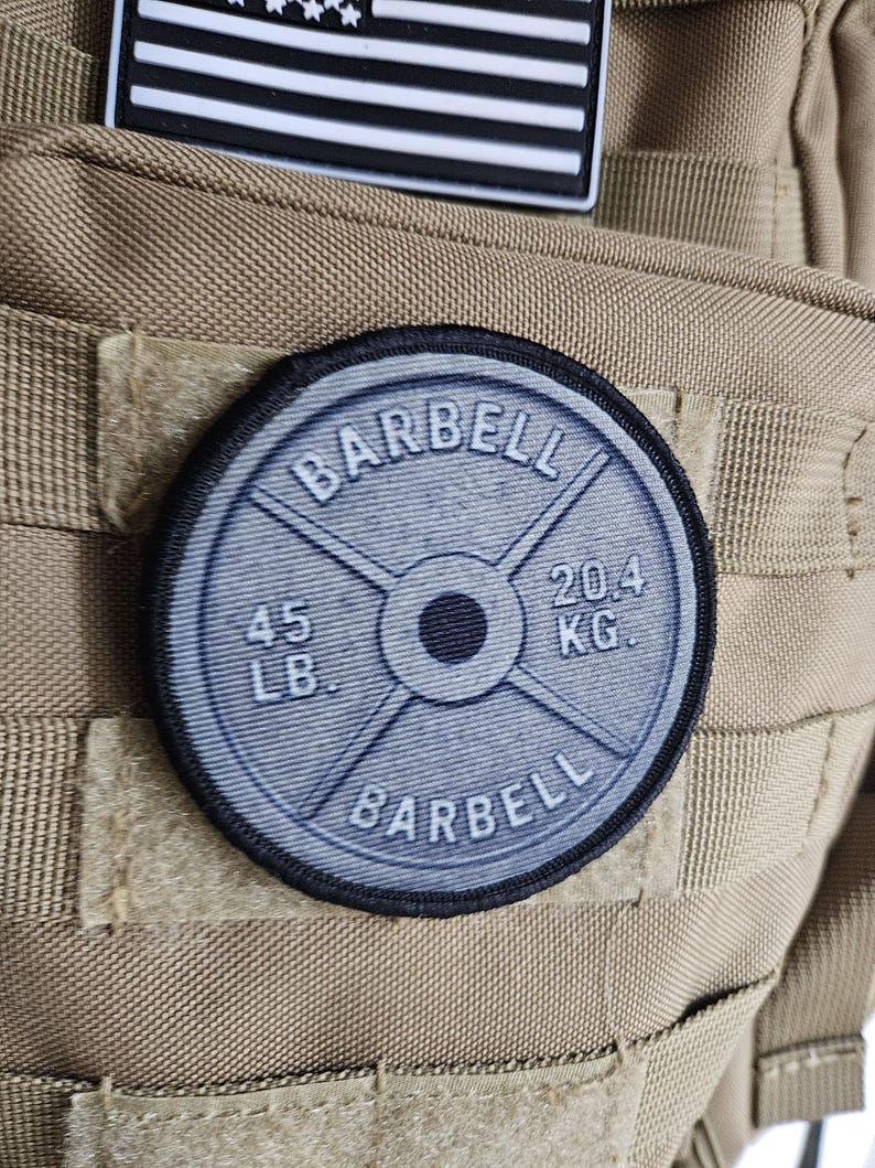 May include: A round, gray barbell weight patch with the word "BARBELL" in a circular pattern. The patch displays "45 LB" and "20.4 KG" on the weight. The patch is attached to a tan tactical backpack, with a black and white American flag patch above it.