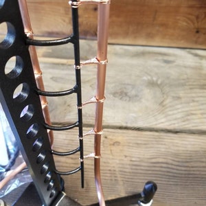 May include: A close-up of a copper pipe with black metal rungs. The pipe is attached to a black metal stand with a wooden base.