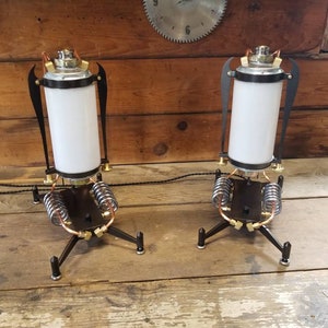 May include: Two steampunk-style table lamps with black metal bases, copper accents, and white glass shades. The lamps have a unique design with springs and gears.