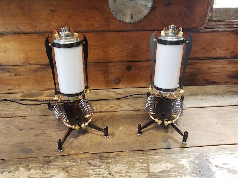 May include: Two steampunk-style table lamps with black metal bases, copper accents, and white glass shades. The lamps have a unique design with springs and gears.