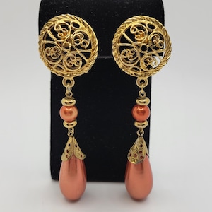May include: A pair of gold filigree drop earrings with orange glass beads. The earrings have a circular top with a delicate design and a teardrop-shaped bottom.