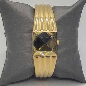 May include: A gold-toned bangle watch with a square face and a clear, faceted crystal. The watch is on a gray pillow.