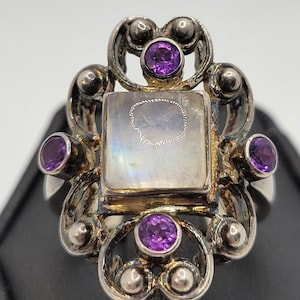 May include: A silver ring featuring a central square moonstone cabochon surrounded by four small round purple amethyst gemstones. The metal setting displays an intricate, ornate scrollwork design with polished silver spheres throughout the frame.