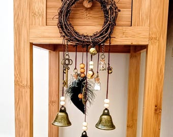 May include: A circular grapevine wreath adorned with hanging charms including three brass-toned bells, a metal skeleton key, two small glass vials with cork stoppers, wooden beads, and a dark feather, all suspended by thin brown cords.